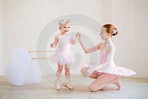 The older sister, a ballerina in a pink tutu and pointe shoes, shows the baby how to practice at the barre