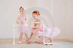 The older sister, a ballerina in a pink tutu and pointe shoes, shows the baby how to practice at the barre