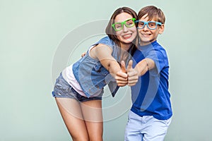 Older sister and her brother with freckles, posing over blue background together, looking at camera with toothy smile and thumbs