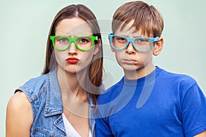 Older sister and her brother with freckles, posing over light blue background together, looking at camera with unhappy faces.