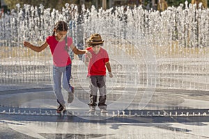 The older sister holds her brother`s hand as they jump over the water jets in the fountain
