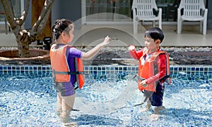 The older sister play rock-paper-scissors game with her younger brother in the swimming pool. Both wear orange life jacket