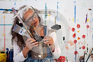 Older woman, artist, in her fifties with grey hair and black glasses and many paintbrushes, copy space