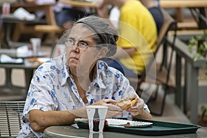 Older woman eating a hamburger at an outside table looking up with an unhappy expression on her face