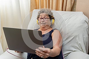 Older woman with headphones using a laptop while sitting on the sofa at home.