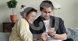 Older woman her grownup son sit on sofa use smartphone