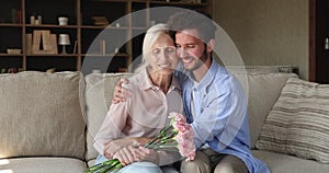 Older woman holds flowers sit on sofa with grownup son
