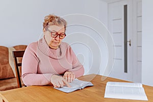 An older, overweight woman reading a book with glasses on while sitting comfortably at home.