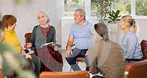 Older woman presides over support group meeting with mature people listening