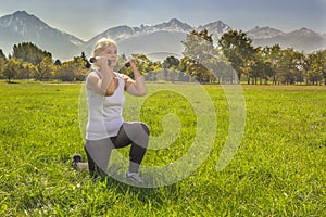 Older woman squats with dumbbells while doing fitness in the park against the backdrop of mountains