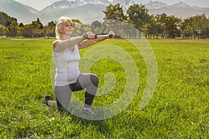 Older woman squats with dumbbells while doing fitness in the park against the backdrop of mountains