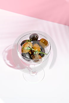 Olive Kalamata in cocktail glass. Black and green olives food on white table with pink wall. Day sunlight with hard shadow of fern