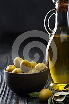 Olive oil in a glass bottle, fresh green olives in a ceramic bowl and olive branch on an old wooden table