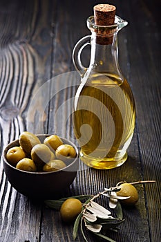 Olive oil in a glass bottle, fresh green olives in a ceramic bowl and olive branch on an old wooden table