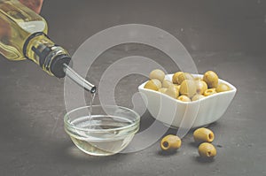 Olive oil pouring from bottle in bowl and fresh olives in ceramic plate and on dark stone table