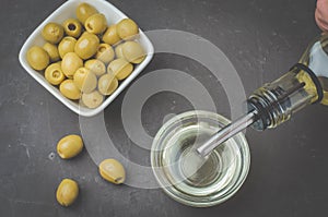 Olive oil pouring from bottle in bowl and fresh olives in ceramic plate and on dark stone table. Top view