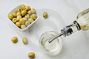 Olive oil pouring from bottle in bowl and fresh olives in ceramic plate and on white stone background. Top view