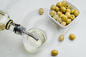 Olive oil pouring from bottle in bowl and fresh olives in ceramic plate and on white stone background. Top view
