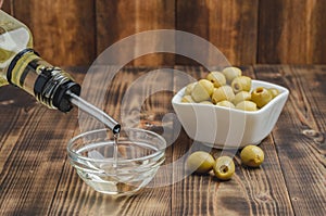 Olive oil pouring from bottle in bowl and fresh olives in ceramic plate and on wooden background