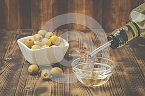 Olive oil pouring from bottle in bowl and fresh olives in ceramic plate and on wooden background