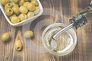 Olive oil pouring from bottle in bowl and fresh olives in ceramic plate and on wooden background. Top view