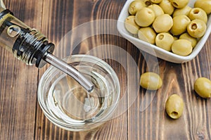 Olive oil pouring from bottle in bowl and fresh olives in ceramic plate and on wooden background. Top view