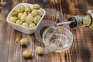 Olive oil pouring from bottle in bowl and fresh olives in ceramic plate and on wooden background. Top view