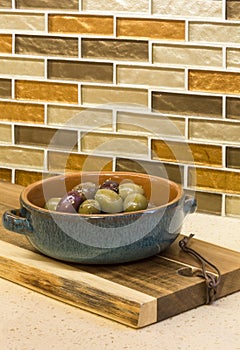 Olives in bowl and wooden cutting board on engineered stone countertop with glass mosaic tile backsplash in home kitchen