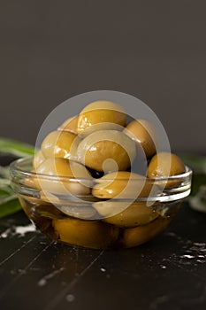 Olives in an extra virgin olive oil placed in a glass bowl and green branches on a dark table with copy space, vertical shot