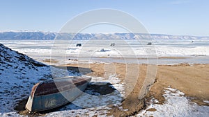 Olkhon island in winter: on the shore is an inverted old dilapidated boat on the ice in the distance go three cars