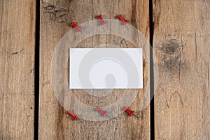One blank business card and red push pins against a wooden background. A white paper rectangle and six push pins lie on top of old