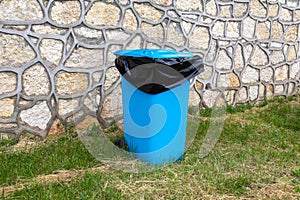 A blue plastic trash can with a black plastic bag near a decorative stone wall