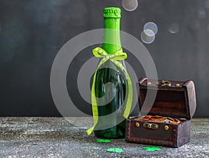 One bottle of green beer with an open chest, coins and three paper-cut clovers on a dark gray cement.