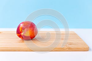 One bright ripe red yellow nectarine lies on a light wood cutting board, white table, sky blue background. Finished product, healt