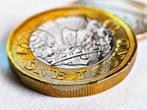 One British pound coin lies on a light white textured surface. Focus is on denomination of coin and name of currency of UK.