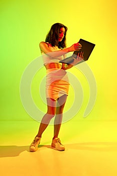 One brunette, charming girl, woman holding laptop and looking at the screen over acid gradient background. Remote work