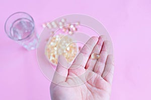 One capsule of omega three on the female palm on a pink background with a glass of water and fish oil top view