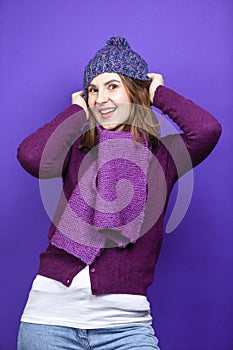 One Caucasian Adult Female Woman In Warm Knitted Hat and Violet Scarf with Lifted Hands Pulling Hat Down While Smiling On Blue