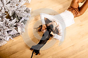 One cheerful girl in a white knitted sweater and brown pants. A curly woman lies on the floor with a black dachshund dog