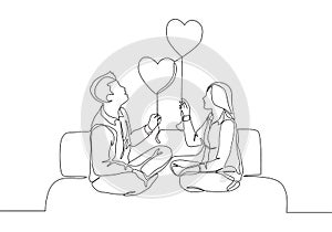 One continuous line drawing of young happy man and woman couple sitting on the couch and holding heart shape balloon together