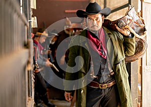 One cowboy hold saddle and look at camera while other two man check the gun in background of stable