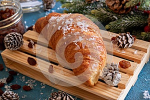 One croissant lies on a wooden board on a blue background. Nearby lies a spruce branch with cones and berries, and there is an ope