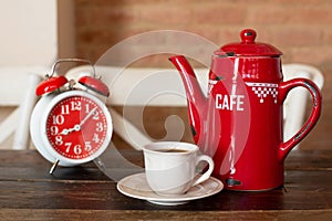 One cup of black coffee, a red coffee pot and an alarm clock of red color in a retro style are on a wooden kitchen table.