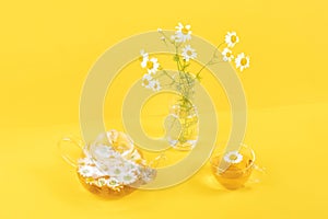 One cup of camomile tea, transparent teapot and vase with daisy-like flowers on yellow background. Chamomile Tea