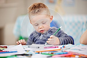 One cute little caucasian boy drawing a picture in a book while sitting alone at table and colouring at pre-school or
