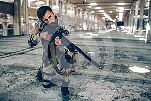 One dark-haired man is standing in long hangar by himself. He has put one knee down on the ground and holding rifle in