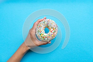 One donut with white icing. An overhead view of a doughnut with colored sprinkles. Female hand holding donut over blue