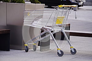 One empty metal shopping cart, with yellow plastic parts, is left on sidewalk outside the store poor people