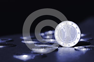 One euro coin standing on edge shining in spotlight against many different coins on black. Money flow