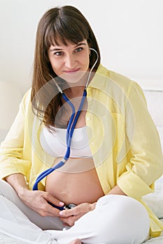 One european pregnant woman with smile is listening heart beat of unborn child inside her tummy by stethoscope while she